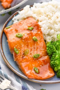 Teriyaki Salmon garnished with green onions and sesame seeds, served on a plate with white rice and broccoli. Fork beside.