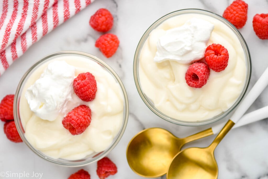 Overhead view of two bowls of Pudding garnished with raspberries and whipped cream, raspberries and spoons beside.